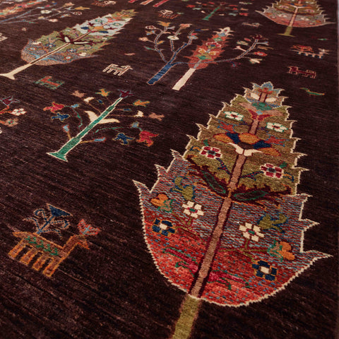 A close-up of a dark brown, hand-knotted Afghan rug with stylised tree motifs in red, green, gold, beige, and white, featuring small geometric and floral details throughout.